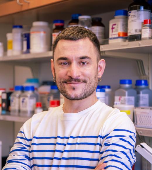 Person in a striped shirt stands smiling with arms crossed in a lab, shelves of chemical bottles visible behind.