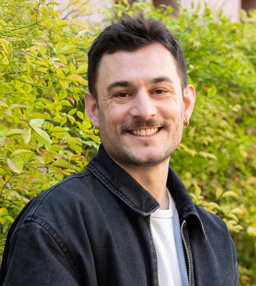 A smiling person with short dark hair, a mustache, and gold hoop earrings is outdoors in front of green foliage. They are wearing a black denim jacket over a white t-shirt. The background consists of lush green leaves, slightly blurred.