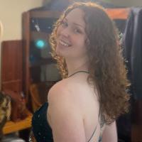 Smiling person with long curly hair wearing a dark strappy dress, turned slightly to the side to show a tattoo on their back, with soft indoor lighting and blurred background elements.