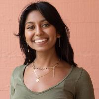 Smiling young woman wearing a green top and gold jewelry, standing in front of a peach-colored wall.