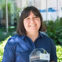 A smiling woman stands outdoors holding a clear glass award. She wears a navy blue shirt and has shoulder-length dark hair with bangs.