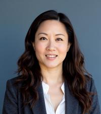 Smiling woman with long, wavy hair, wearing a gray blazer and white shirt, against a blue background.