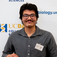 Smiling young man with glasses and mustache wearing a name badge in front of UC Davis banner.