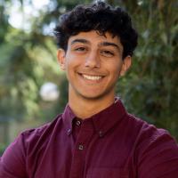 A UC Davis student smiles confidently outdoors with arms crossed in front of a background of greenery. He has short curly hair and is wearing a maroon button-up shirt.