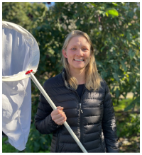 A smiling person in a black puffer jacket holds a white insect net, standing outdoors in front of leafy green trees on a sunny day.