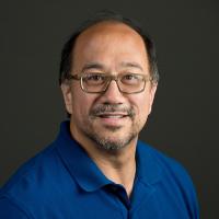 Headshot of a middle-aged man wearing glasses and a royal blue polo shirt, smiling in front of a dark gray background.