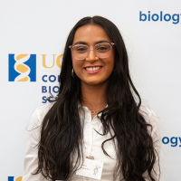 Smiling woman with long dark hair and glasses, white blouse, conference logo backdrop