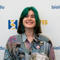 Smiling person with green hair and patterned shirt in front of logo-covered backdrop