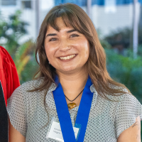Smiling woman with shoulder-length brown hair wearing blue medal ribbon and name badge outdoors