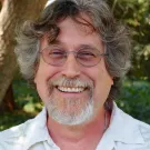 Smiling older man with gray hair, beard and glasses wearing a white shirt among trees