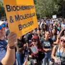 A person in sunglasses and a cap holds a yellow "Biochemistry & Molecular Biology" sign at a university orientation. Behind them, a diverse group of students watches attentively, many wearing lanyards, while trees and clear skies frame the outdoor event.