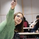 A professor in a green sweater points at a screen while a student in a black cap listens intently; other students take notes in the background.