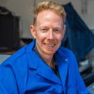 Smiling man with blond hair wearing a blue lab coat in a workshop