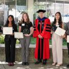 Six people stand in a row outdoors in front of a glass-walled building. Five students, holding awards and certificates, pose on either side of a man in a red and black academic robe and cap. The students appear to be honored at a recognition event.