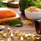 A close-up of nutrient-rich foods and supplements on a wooden surface, featuring a raw salmon fillet on a cutting board, a bowl of sesame seeds, fresh spinach leaves, broccoli, and a white spoon containing olive oil and an olive. Nearby, an amber bottle is tipped over, spilling golden omega-3 capsules.