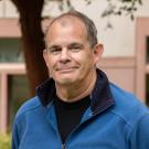 Portrait of a man with short gray hair, wearing a blue fleece jacket over a black shirt, standing outdoors with a building and trees in the background. He has a slight smile and is looking directly at the camera.