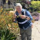 A person wearing a cap and field research vest examines orange flowers in a vibrant, well-maintained residential garden. They are surrounded by blooming plants and shrubs along a sidewalk on a sunny day.
