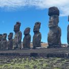  A row of large stone moai statues standing on a stone platform (ahu) at Ahu Tongariki on Easter Island. The statues face inland, with one wearing a topknot-like stone hat (pukao). The background shows grassy hills and a bright blue sky with scattered clouds.
