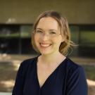 Headshot photograph of smiling woman with glasses and navy blouse, blurred greenery