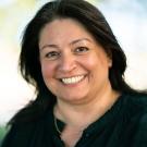 The image features an individual with long dark hair wearing a black top and a gold necklace. The background appears to be outdoors with blurred greenery, suggesting a shallow depth of field typically used in portrait photography.