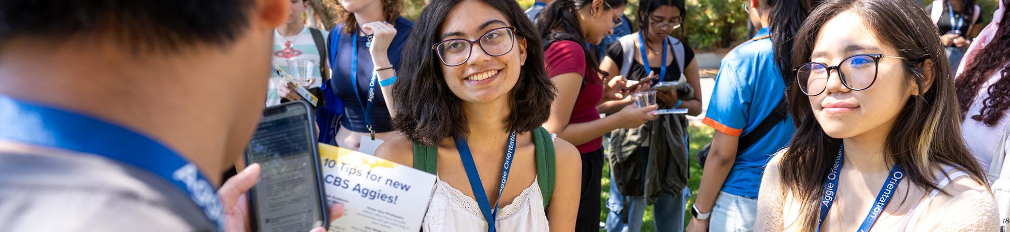 A group of UC Davis students wearing Aggie Orientation lanyards gathers outdoors during a sunny event. One student in the center smiles while holding a "10 Tips for New CBS Aggies!" handout. Others are engaged with their phones or talking in the background, creating a lively and welcoming atmosphere.