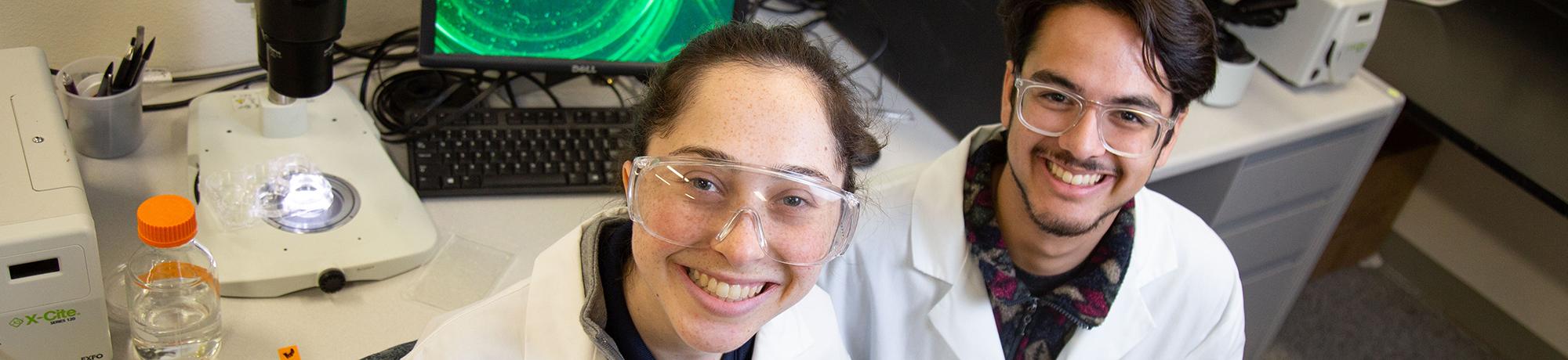 Two smiling students in lab coats and safety goggles working in a lab, with microscopes and a computer displaying a green image in the background.