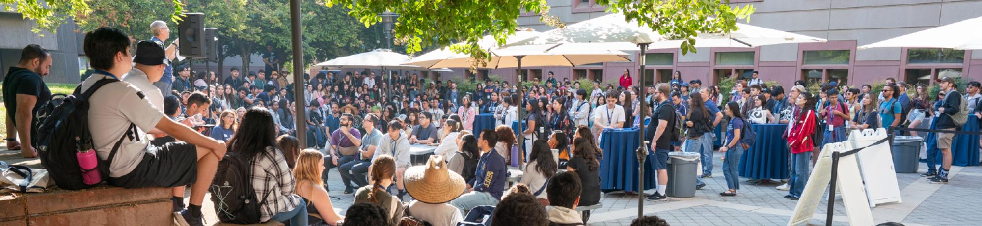 A large group of students gathers outdoors under umbrellas for the college’s annual welcome event and opening remarks.