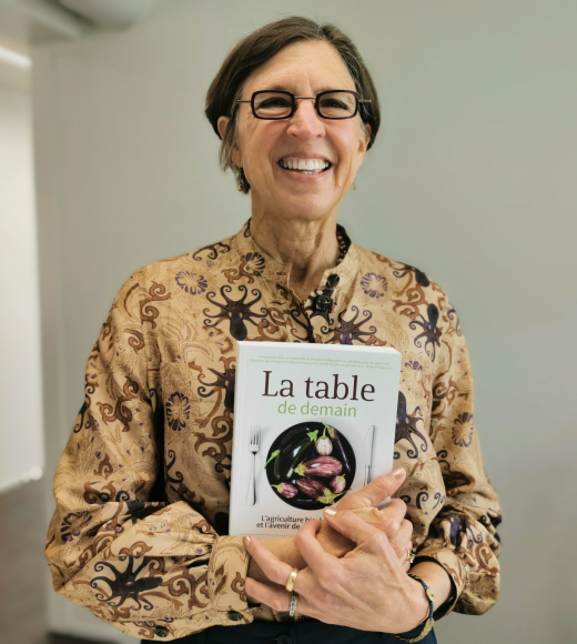 The image depicts Dr. Pamela Ronald holding a copy of La Table De Demain